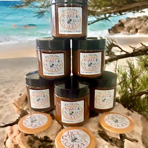 以下が含まれることがあります： 「Bee Eye DCW Lucky」と「All Natural」のラベルが付いた、黒い蓋の小さな茶色の瓶が積み重ねられています。「Bee Lucky」の文字が入った丸いオレンジ色のアイテムもいくつか見えます。背景には、海と緑のあるビーチの風景が写っています。