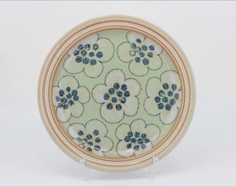 May include: A round ceramic plate featuring a light green background decorated with a repeating pattern of white flowers with dark blue centers. The plate has a raised rim with thin, concentric orange-brown circular lines along the edge.