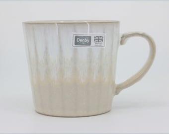 May include: A ceramic mug with a vertical ribbed texture and a two-tone glaze, featuring a white upper section that transitions into a light beige base. A rectangular sticker on the front displays the text Denby 1809 and a Union Jack flag icon.