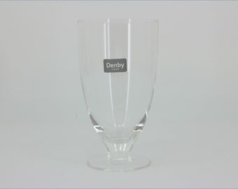 May include: A clear glass goblet with a smooth, curved bowl and a short, sturdy stem resting on a circular base. The glass features a rectangular sticker on the front with the white text Denby 1809 printed on a dark gray background.