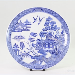 May include: A round, white ceramic plate with a blue Willow pattern design. The plate features a detailed landscape scene with pagodas, bridges, and birds. The rim has a decorative blue border.
