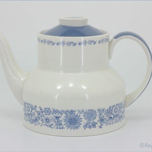 Op de afbeelding: Witte keramische theepot met een blauw deksel en handvat. De theepot heeft een blauw bloemmotief rond de basis en de bovenkant. De tuit is gebogen en het handvat is een simpele lus.
