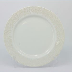 Op de afbeelding: Een witte keramische plaat met een decoratief, lichtbeige bloemmotief rond de rand. De plaat is rond en staat op een doorzichtige plastic standaard. Deze plaat is geschikt voor het serveren van voedsel.