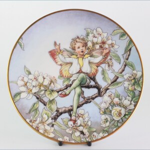 Puede incluir: Un plato de porcelana con un borde dorado presenta una pintura de un hada con alas de mariposa sentada en una rama de un árbol floreciente. El hada lleva una camisa blanca, pantalones verdes y zapatos rojos. Las flores son blancas y el fondo es azul claro.
