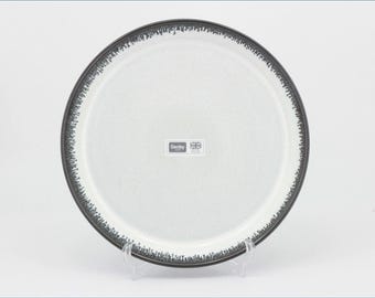 May include: A round ceramic dinner plate with a speckled light gray center and a dark charcoal rim featuring a textured, drip-like pattern. A rectangular white label in the center displays the text Denby and a small Union Jack flag icon.
