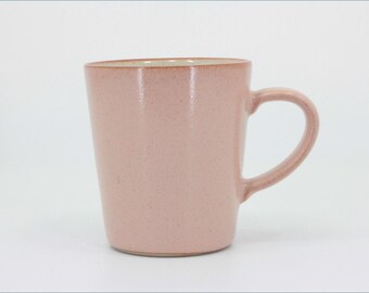 May include: A light pink ceramic mug with a speckled finish and a smooth, cylindrical body. The mug features a curved handle on the right side and a wide, open top rim, showcasing a simple, functional design with a consistent pastel color tone.