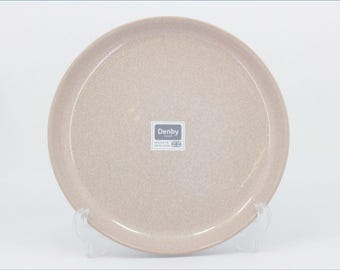 May include: A round, light beige ceramic plate with a speckled finish. The center of the plate features a square sticker with the text Denby 1809 and Made in England with a small Union Jack flag icon. The plate has a smooth, glossy surface.