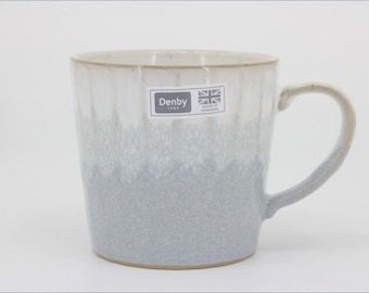 May include: A ceramic mug featuring a two-tone glaze with a creamy white upper section and a textured light gray base. A rectangular sticker on the front displays the text Denby 1809 and a Union Jack flag with the words Made in England.