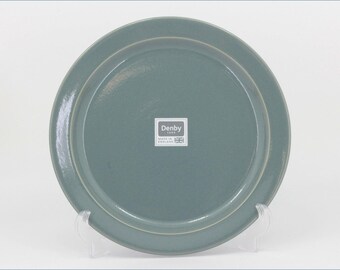 May include: A round, sage green ceramic plate with a smooth, glossy finish. Centered on the plate is a white square sticker featuring the text Denby 1809, Made in England, and a small graphic of the Union Jack flag. The plate has a raised rim.
