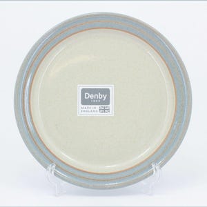 Puede incluir: Un plato redondo de cerámica con un centro beige claro y un borde ancho de color azul grisáceo moteado con una fina línea de acento marrón. Una pegatina cuadrada en el centro muestra el texto Denby 1809 Made in England con una bandera británica.