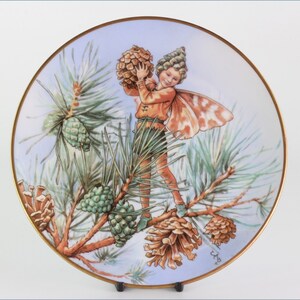 Può includere: Un piatto in porcellana con un bordo dorato presenta un'illustrazione fantasiosa di una fata che tiene un pigna. La fata indossa una tunica marrone e pantaloni verdi, e ha un'ala di farfalla. Il piatto è decorato con uno sfondo blu e bianco, e un ramo di pino con pigne.