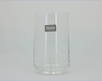 May include: A clear glass tumbler with a cylindrical shape and a thick base. A rectangular grey sticker is applied to the front surface, featuring the white text Denby 1809. The glass has a smooth, transparent finish and a simple, functional design.