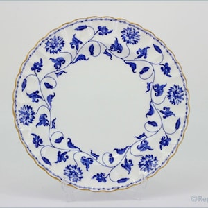 May include: White ceramic plate with a blue floral design and a gold rim. The plate has a scalloped edge and a central floral pattern. The design includes flowers and vines. The plate is displayed on a clear stand.