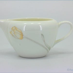 May include: Cream-coloured creamer with a tulip design. The creamer has a spout and a handle. The tulip is yellow and orange with a grey stem and leaf. The creamer is made of ceramic.