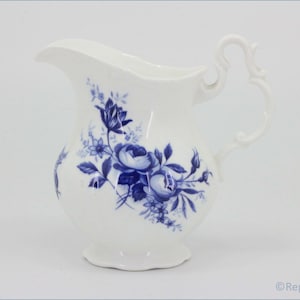 Op de afbeelding: Witte keramische kan met een blauw bloemmotief. De kan heeft een gebogen tuit en een sierlijk handvat. Het bloemmotief omvat rozen en andere bloemen. Het is waarschijnlijk bedoeld voor het serveren van drankjes.