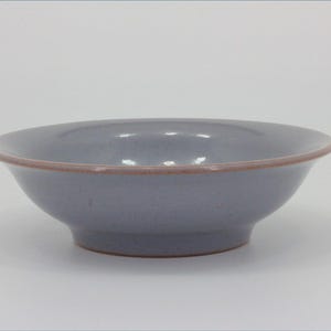 Puede incluir: Un cuenco de cerámica redondo y poco profundo con un esmalte azul bígaro claro y un acabado moteado. El cuenco presenta un borde distintivo ligeramente acampanado con un borde fino de color terracota expuesto que contrasta con la superficie.