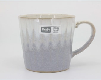 May include: A ceramic mug featuring a two-tone glaze with a creamy white top transitioning into a soft lavender-grey base. A rectangular sticker on the front displays the text Denby 1809 and Made in England with a small Union Jack flag icon.
