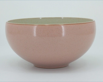 May include: A round ceramic bowl with a smooth, dusty pink exterior featuring a subtle speckled texture. The interior of the bowl is a contrasting light cream color with a similar speckled finish, showing a clean, curved rim and base.