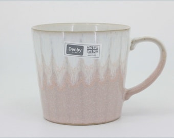 May include: A ceramic mug featuring a white upper section that transitions into a dusty pink base with a subtle vertical drip pattern. A rectangular sticker on the front displays the text Denby 1809 alongside a small Union Jack and Made in England.