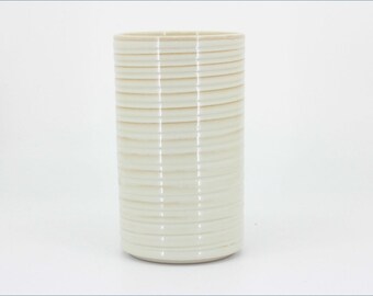 May include: A cylindrical ceramic vase with a glossy off-white finish. The exterior surface features a series of evenly spaced, horizontal ridges that wrap around the entire body of the vessel, creating a subtle texture from top to bottom.
