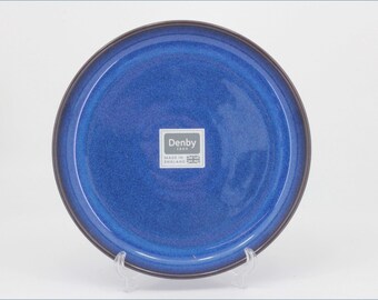 May include: A round ceramic plate with a deep blue glazed finish and a dark brown rim. The center features a white square sticker with the text Denby 1809 Made in England and a small Union Jack flag icon printed in dark gray.