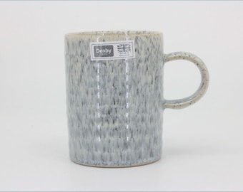 May include: A cylindrical ceramic mug with a light gray and white speckled glaze finish. The mug features a side handle and a rectangular sticker near the rim that reads Denby 1809 with a Union Jack flag and the text Made in England.