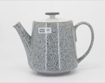 May include: A ceramic teapot with a speckled gray and white finish, featuring a tapered body, a curved handle, and a spout. The lid has a dark gray knob. A rectangular label on the front displays the text Denby and a small Union Jack flag icon.