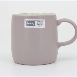 May include: A matte mauve ceramic mug with a rounded body and a curved handle. A rectangular sticker on the front features the text Denby 1809, a small Union Jack flag icon, and the words Made in England in white text on a dark gray background.
