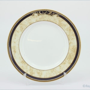 Puede incluir: Un plato de cena de cerámica blanca con un borde dorado y azul. El borde tiene un diseño de cuerda y un patrón floral.