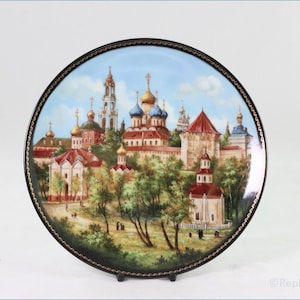 Könnte beinhalten: Ein runder Porzellanteller mit schwarzem Rand und goldener Verzierung zeigt eine handgemalte Darstellung eines russisch-orthodoxen Klosters mit mehreren Gebäuden, blauem Himmel und grünen Bäumen.
