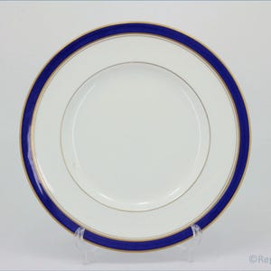 Puede incluir: Plato de cerámica blanca con borde azul marino y fino acento dorado. El plato tiene un diseño sencillo y elegante, adecuado para cenas formales. El plato se exhibe sobre un soporte transparente.