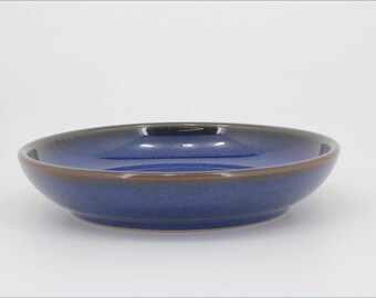 May include: A round ceramic bowl with a deep blue interior and exterior glaze. The rim of the bowl features a distinct brown band that contrasts with the blue finish. The bowl has a smooth, curved silhouette and a shallow depth, resting on a base.