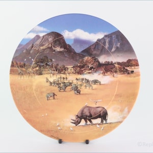 Könnte beinhalten: Ein runder Keramikteller mit einem farbenfrohen Bild einer Savannenlandschaft mit einem schwarzen Nashorn, Zebras, Giraffen und Vögeln. Der Teller hat einen blauen Himmel mit weißen Wolken und braunen Bergen im Hintergrund.