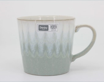 May include: A ceramic mug featuring a two-tone glaze with a white upper section and a sage green lower section. The surface displays a subtle scalloped pattern where the colors meet. A rectangular sticker reads Denby 1809 Made in England.