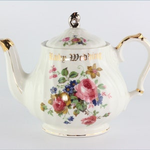 Könnte beinhalten: Weiße Teekanne mit Blumenmuster und goldenen Verzierungen. Die Teekanne hat einen Deckel und einen Henkel. Der Text "Baby Wedding" ist in Gold auf der Seite der Teekanne geschrieben.