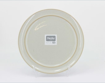 May include: A round, light beige ceramic plate with a smooth, matte finish and a subtle tan rim. The center features a square sticker with the text Denby 1809 and Made in England, accompanied by a small graphic of the Union Jack flag.