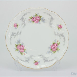 Puede incluir: Un plato de porcelana blanca con borde festoneado y ribete dorado. El plato presenta ramos de rosas rosas y detalles decorativos grises. El plato se exhibe sobre un soporte transparente.