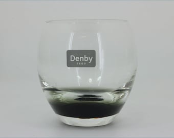 May include: A clear glass tumbler with a rounded, bulbous shape. The base features a dark, smoky gray tint that fades into the transparent upper section. A rectangular gray label is printed on the front with the white text Denby 1809.