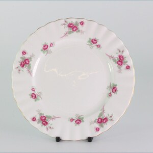 Peut inclure: Assiette en céramique blanche avec un bord festonné et un bord doré. L'assiette est décorée de motifs floraux de roses roses et de feuilles vertes. L'assiette est présentée sur un support noir sur fond blanc.