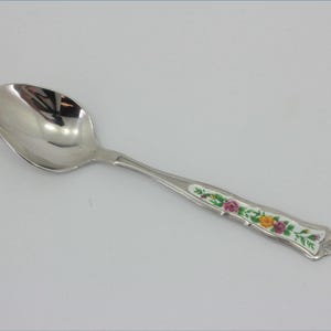 May include: A silver-toned metal spoon featuring a polished, reflective bowl and a decorative handle. The handle is inlaid with a white panel displaying a colourful floral pattern of pink and yellow roses with green leaves and stems.