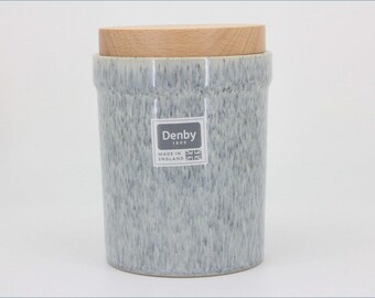 May include: A cylindrical ceramic storage canister with a speckled light blue and white glaze. The container features a flat, light-colored wooden lid and a square label on the front that reads Denby 1809 Made in England with a small flag icon.