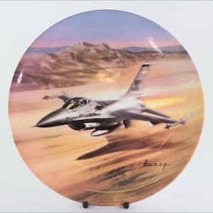 Könnte beinhalten: Ein Keramikteller mit einem Gemälde eines grauen Kampfjets, der über eine Wüstenlandschaft fliegt. Der Jet trägt die Buchstaben "YP 1426" an der Seite. Der Teller hat einen goldenen Rand und eine Signatur in der rechten unteren Ecke.