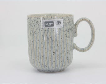 May include: A ceramic mug with a speckled light blue and cream finish featuring a vertical ribbed texture. A rectangular sticker on the front displays the text Denby 1809 and a Union Jack flag with the words Made in England printed below it.