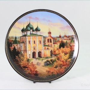 Könnte beinhalten: Ein runder Keramikteller mit einem gemalten Bild einer russisch-orthodoxen Kirche mit grünen Zwiebelkuppeln und einer gelben Fassade. Die Kirche ist von Bäumen und einem Weg zum Eingang umgeben. Der Teller hat einen schwarzen und goldenen Rand.