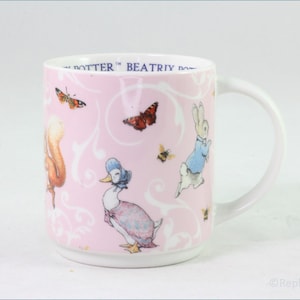 May include: A pink ceramic mug with a white handle features a whimsical design of Peter Rabbit, a duck, a squirrel, and butterflies. The mug has the text "Beatrix Potter" on the side.