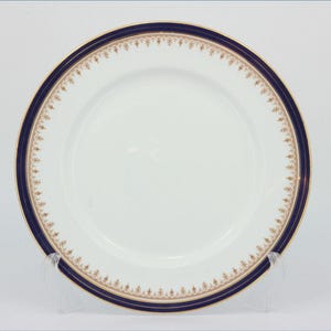 Può includere: Piatto in ceramica bianca con bordo blu navy e oro. Il piatto ha una larga fascia blu navy con un motivo floreale dorato e un bordo dorato. Il piatto è esposto su un supporto trasparente.