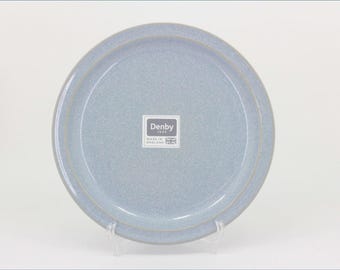 May include: A round, light blue ceramic plate with a speckled finish and a raised rim. A square white sticker is centered on the plate, featuring the text Denby 1809, Made in England, and a small graphic of the Union Jack flag in the bottom corner.