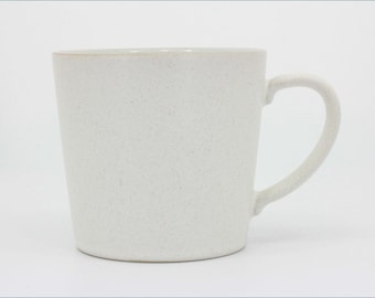 May include: A cylindrical ceramic mug with a speckled off-white finish and a smooth, curved handle on the right side. The mug features a slightly tapered body and a plain, unadorned surface, showcasing a consistent light-colored texture throughout.