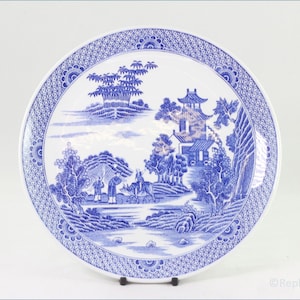 May include: A blue and white ceramic plate with a detailed landscape scene. The plate features a pagoda, trees, figures, and a patterned border. The design is reminiscent of traditional oriental porcelain.