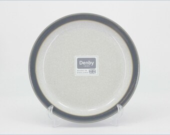 May include: A round ceramic plate with a speckled light gray center and a dark charcoal gray rim. The center features a white square label with the text Denby 1809 Made in England and a small Union Jack flag icon printed in dark gray ink.